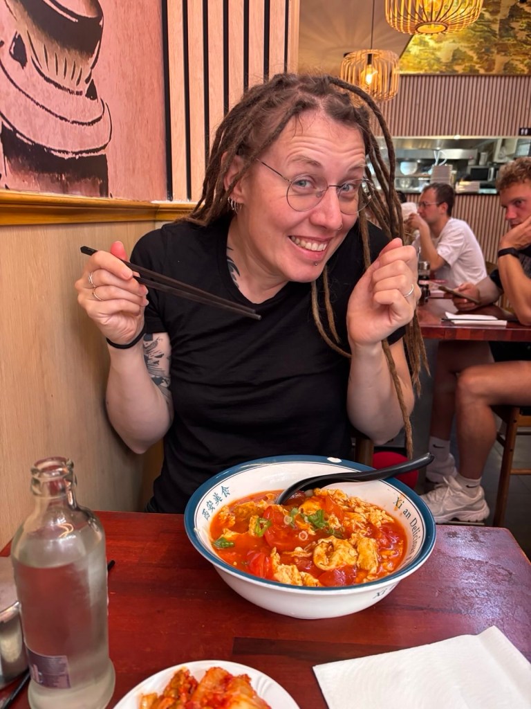 Silke sitzt freudig strahlend mit Stäbchen in der Hand vor einer großen Schüssel chinesischer Nudelsuppe mit Tomate und Ei. Auf dem Teller ist ein Teil des Namens des Restaurants zu lesen: „Xi‘an Delicious …“ Im Hintergrund sitzen weitere Gäste.