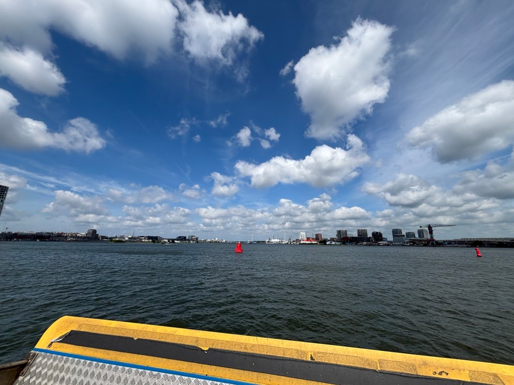 Über die Rehling einer Fähre ist Wasser und im Hintergrund weitere Gebäude Amsterdams zu sehen. Am blauen Himmel zeigen sich einige Wolken.