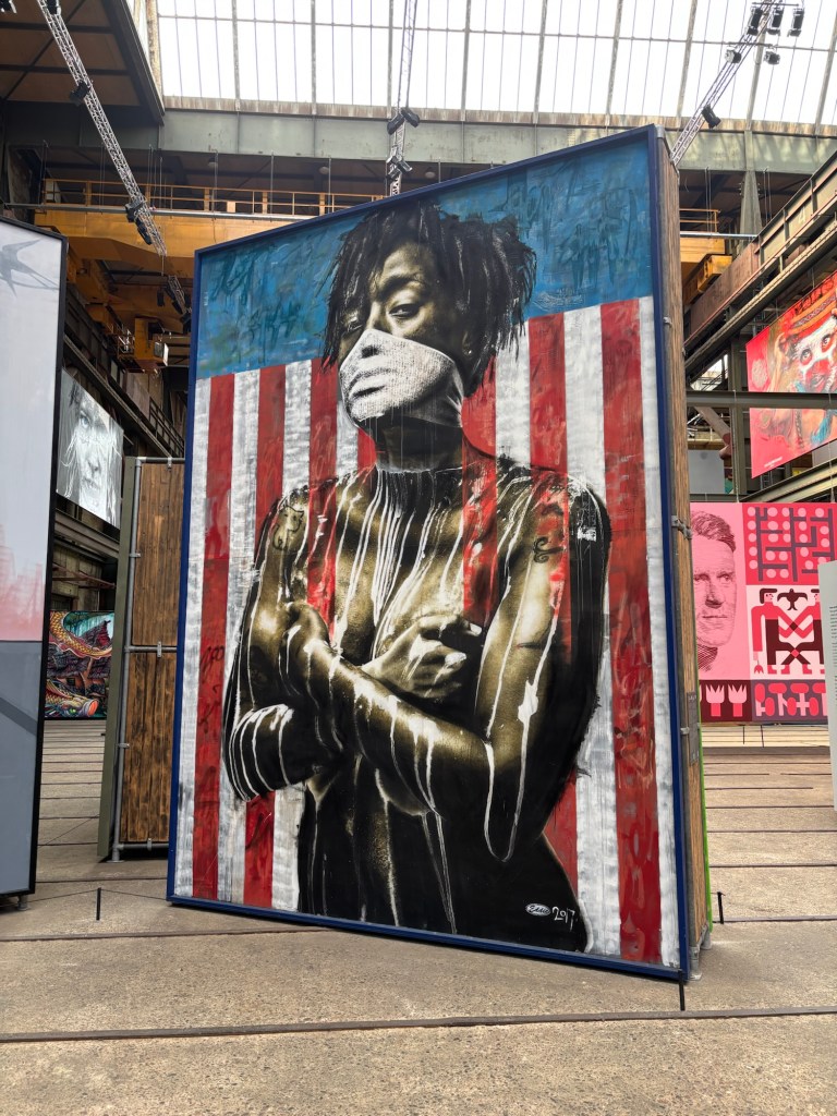 Eine anklagend blickende schwarze weiblich gelesene Person mit einem weißen Tuch vor dem Mund steht vor einer angedeuteten US-amerikanischen Flagge. Ihre Arme sind verkreuzt und bedecken dabei gleichzeitig die nackten Brüste. Den Oberkörper zieren weiße verlaufene Farbflecken.