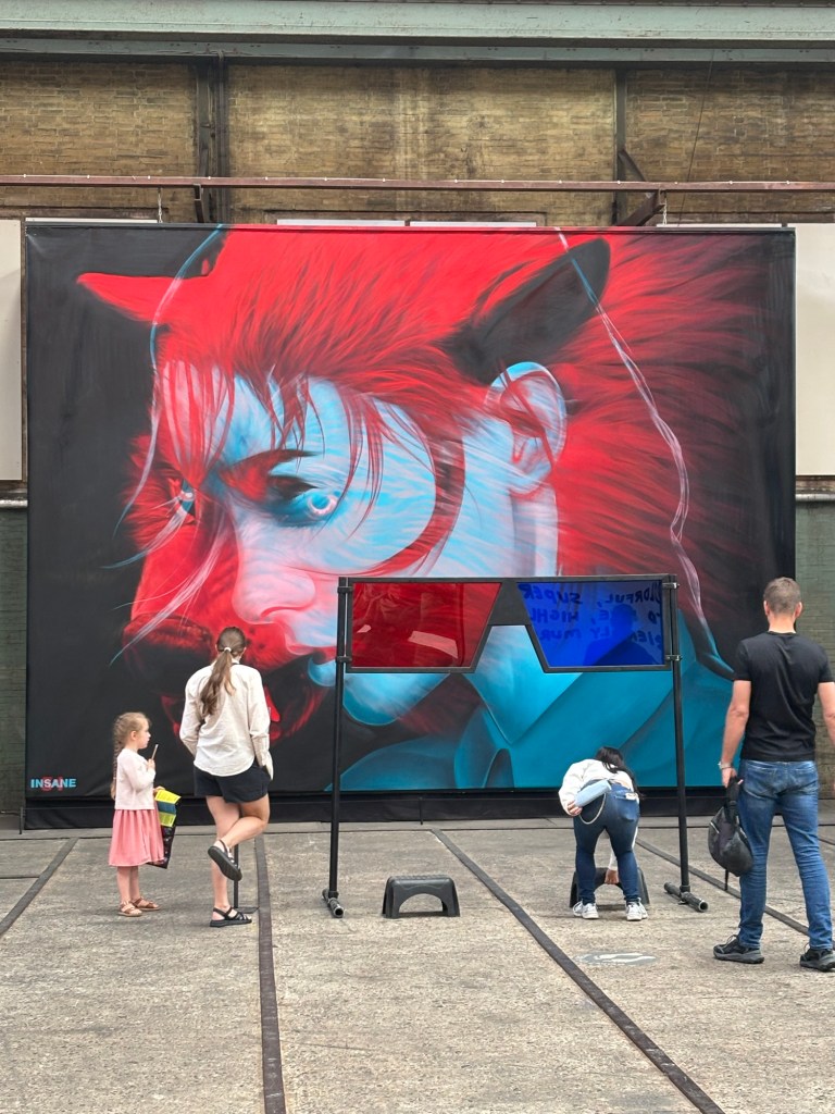 Das Gemälde zeigt das verärgerte Gesicht einer jungen Person in einem Blauton und darüber einen Wolfskopf in rötlichen Farben. Vor dem Gemälde ist eine übergroße Brille platziert. Das linke Glas ist rot eingefärbt, das rechte blau. Vor den Gläsern stehen zwei kleine Hocker. Eine Person schiebt sich einen der Hocker gerade zurecht.