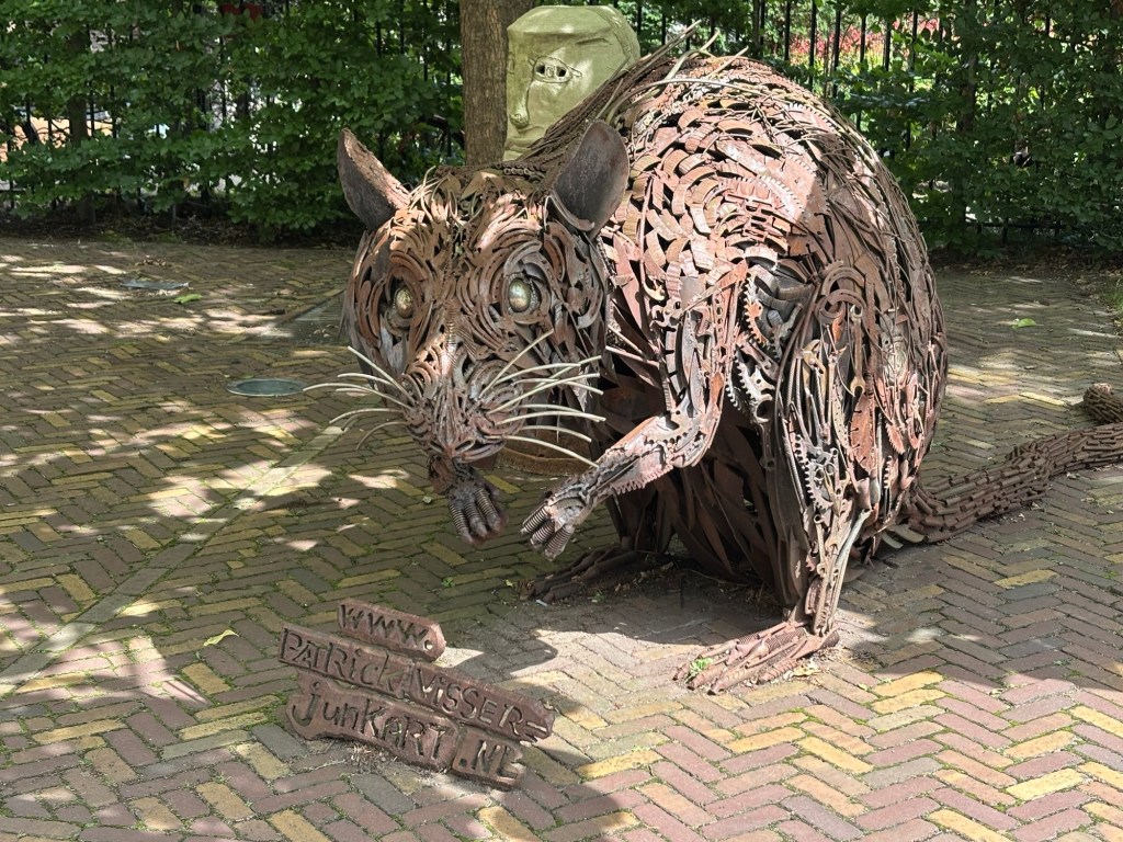 Die Skulptur einer Ratte bestehend aus unterschiedlich geformten Metallteilen. Die Ratte hat ihre Vorderpfoten angehoben und guckt den Betrachter direkt an. Vor ihr weist ein Schild auf die Website des Künstlers hin: junkart.