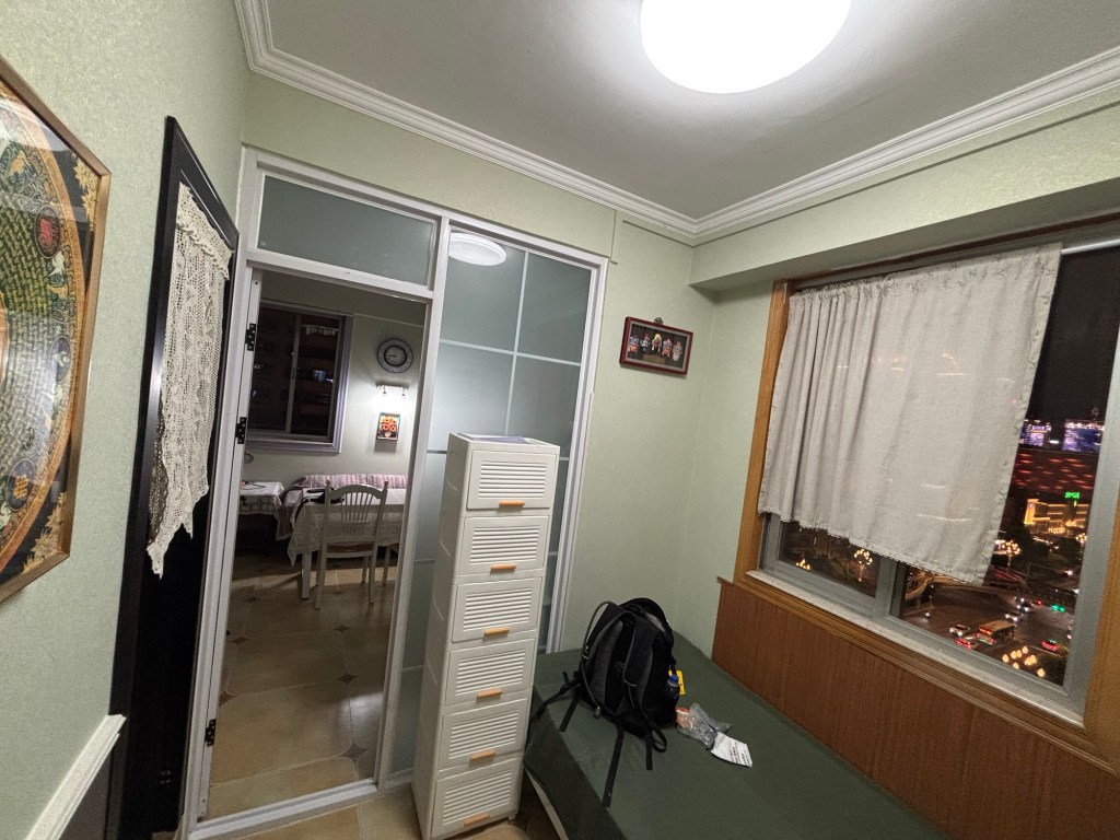 Ein kleiner Raum, rechts ein schmales, abgedecktes Einzelbett, darauf mein kleiner Tagesrucksack. Über dem Bett ein Fenster, links ein Durchgang mit weißen Häkelvorhängen, die die obere Hälfte des Durchgangs verdecken. Geradeaus führt eine Tür in ein weiteres Zimmer.