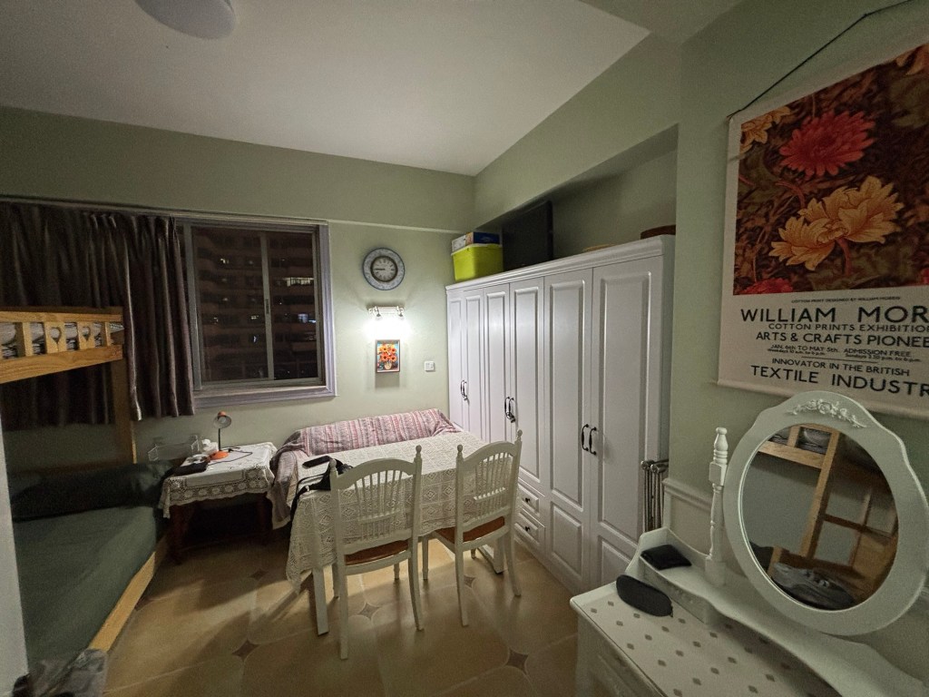 Der Hauptraum mit Tisch, zwei Stühlen und einer Couch. Vorne rechts eine kleine Schminkkommode mit Spiegel. Rechts teilweise vom Tisch verstellt Schränke, nur der vordere ist unverstellt zu öffnen. Die anderen Schranktüren sind mit Vorhängeschlössern gesichert. Links sind Ansätze des Bettes zu erkennen.