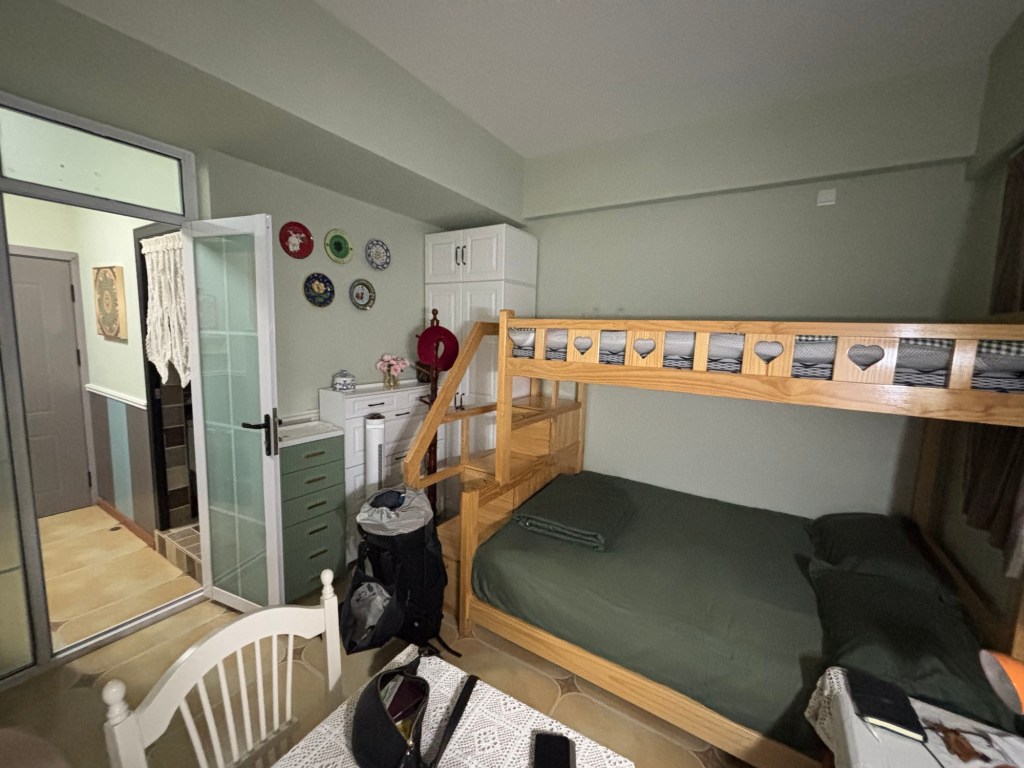 Am unteren Rand die Tischkante mit Häkeldecke, darauf meine Bauchtasche. Ein Stockbett, unten mit Doppelbett ist unten bezogen. Eine kleine Treppe führt zum oberen Bett. Neben dem Bett ein kleiner Tisch. Neben der Treppe zum oberen Teil ein Schrank, ein Garderobenständer und an der Wand eine große und eine etwas kleinere Kommode mit Schubladen. Daneben wieder die Tür, die zum Vorraum und Badezimmer führt.