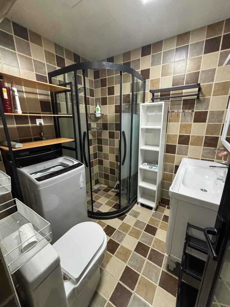 Ein in verschiedenen Brauntönen gefliestes Badezimmer mit abgetrennter Duschkabine. Rechts ein Waschbecken und offenes Regal zum Verstauen von Gegenständen. Links vorne eine Toilette mit Sitz, daneben an der Wand eine graue Toploader Waschmaschine, darüber ein Regal in dem noch Wachmittel und Shampooflaschen stehen.