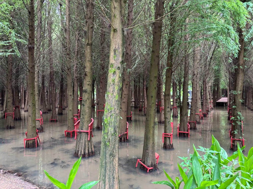 In Reihen gepflanzte Bäume haben in verschiedenen Höhen rote Stühle um ihre Stämme montiert. Die Sitzflächen sind unterbrochen, so dass es wirkt, als seien die Bäume durch die Stühle gewachsen. 