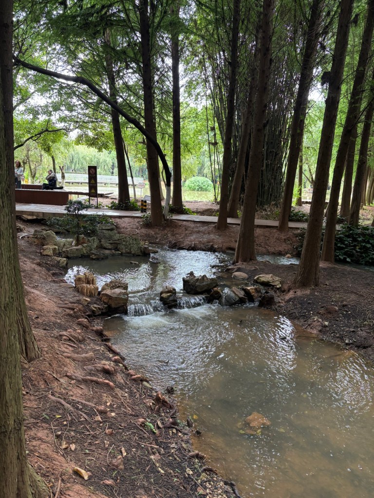 Ein ruhig fließendes Bächlein, gesäumt von Bäumen. Die Erde wirkt lehmig und hat eine rötliche Färbung. Im Hintergrund kreuzt ein gepflasterter Weg. 