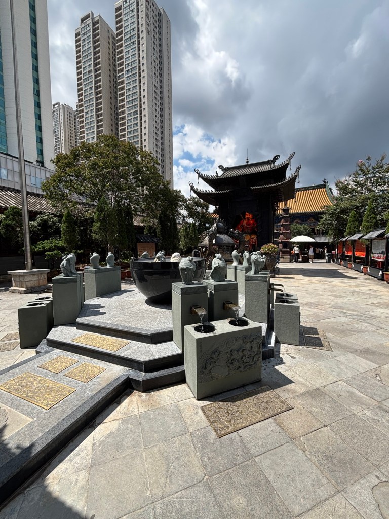 Der gepflasterte Innenhof des Tempels weist eine symmetrische Anordnung mehrerer kleiner Brunnen auf, aus denen Wasser in kleine Schalen fließt. Das Dach des Tempels ist in typisch chinesischer Bauweise an den Ecken nach oben geschwungen. Im Hintergrund sind Hochhäuser zu sehen. 
