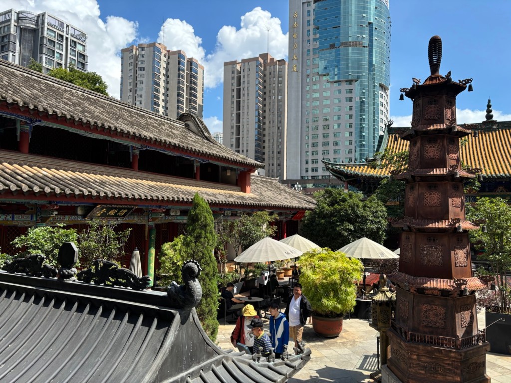 Ein weiterer Innenhof des Tempels mit kleinen Bäumen, Sträuchern und Tischen mit Sonnenschirmen. Die Sonne scheint, die Szene wirkt friedlich. Im Hintergrund bilden Hochhäuser einen Kontrast. 