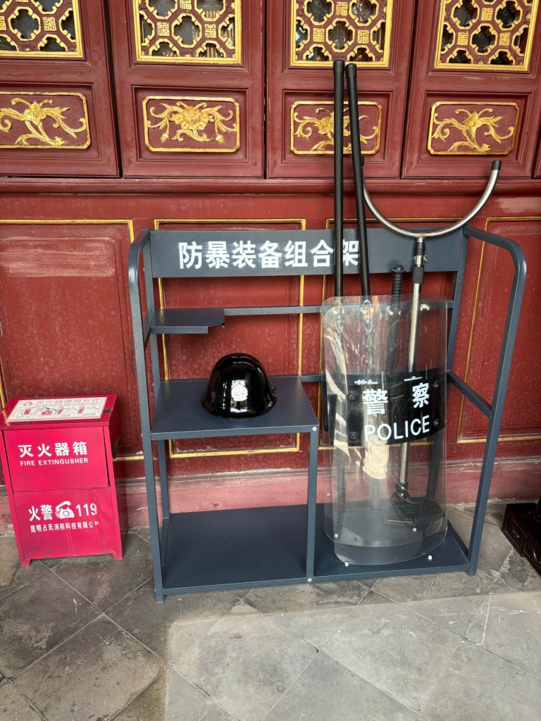 An einer Holzwand im Tempel lehnen Schlagstock, Schild und Helm mit der Aufschrift „police“. Daneben ein Feuerlöscher. 