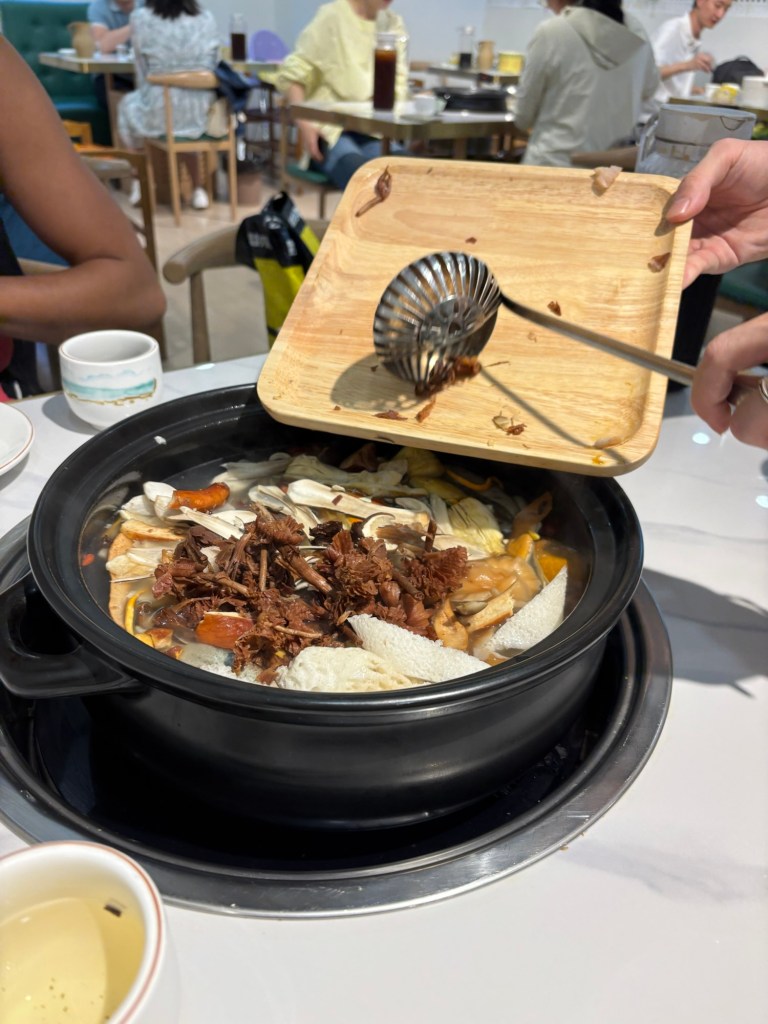 Ein großer Topf steht in der Mitte eines Tisches auf einem Gaskocher. Es wurde gerade ein Tablett mit Pilzen darin ausgeleert.  