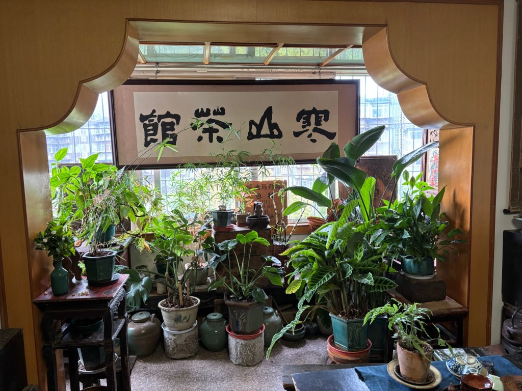 Im Fenster hängt eine Kalligraphie im Querformat. Darum und davor zahlreiche Pflanzen. Der Blick nach draußen ist kaum möglich, dennoch fällt Licht hinein. 