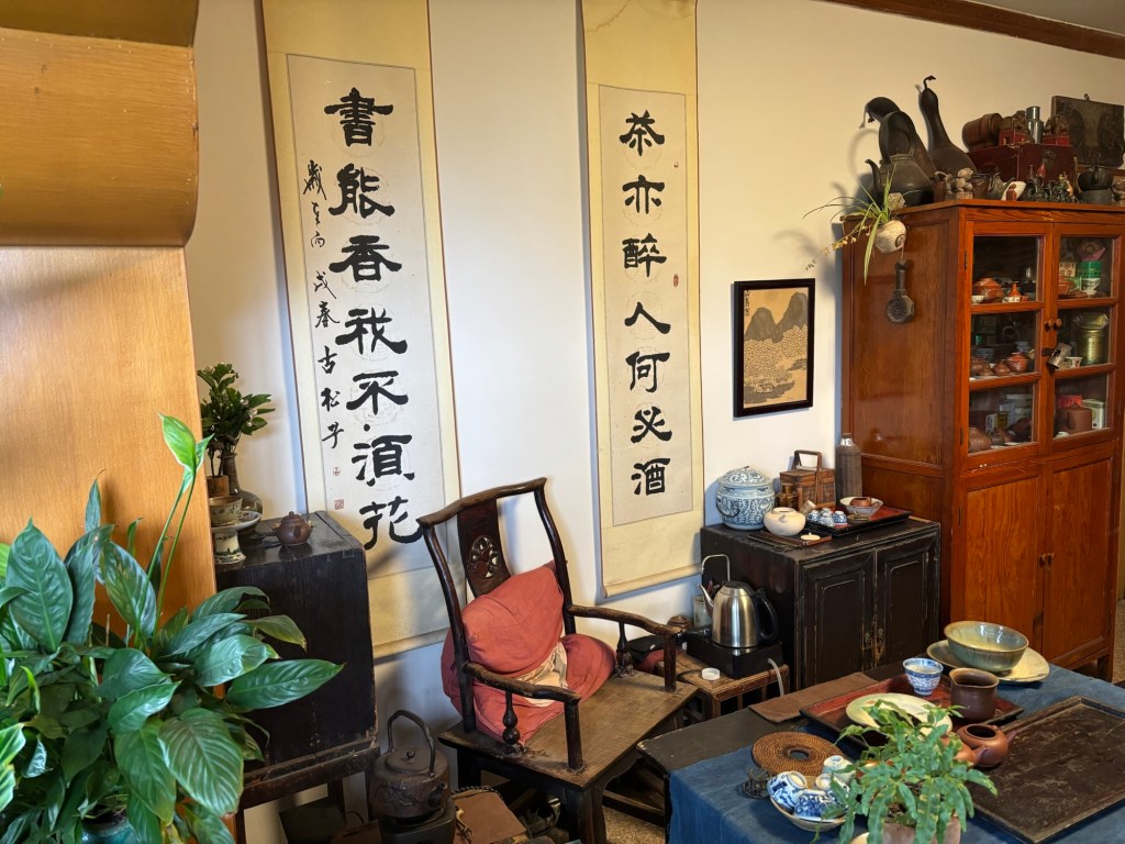 Ein Lehnstuhl, dahinter zwei Kalligraphien längs, ein Wasserkocher und an der dem Stuhl zugewandten Tischkante Teekanne und weiteres Equipment. 