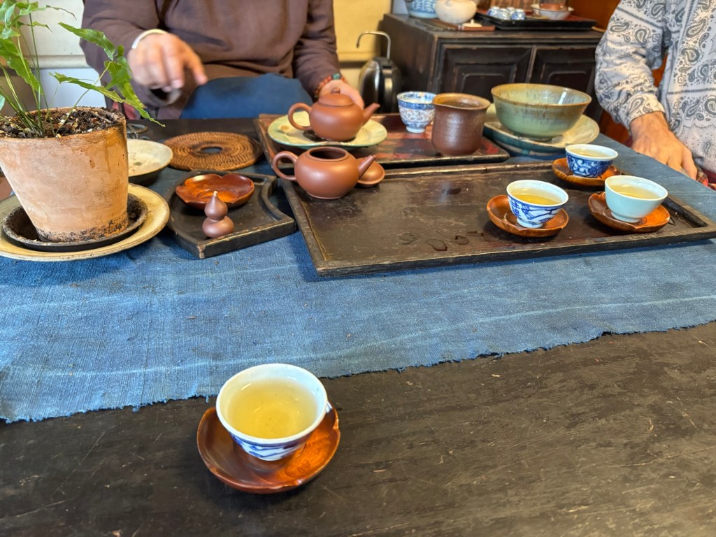 Im Vordergrund eine kleine gefüllte Teetasse. Weitere gefüllte Tassen sind zu sehen. Auf dem Lehnstuhl sitzt eine Person und gestikuliert in Richtung der Teekanne