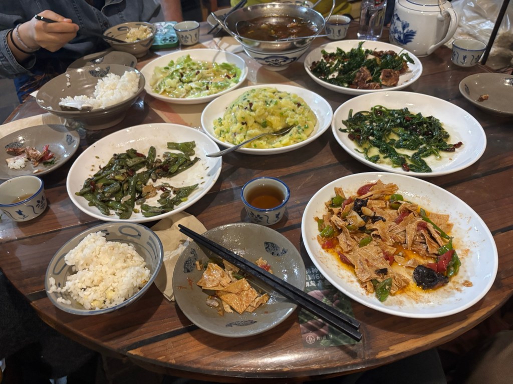 Ein großer runder Tisch mit Gedeck für fünf Personen. In der Mitte stehen sieben verschiedene Gerichte, die in noch unterschiedlicher Quantität vorhanden sind und eine Schüssel Reis. Aus der Perspektive des Tellers der fotografierenden Person sind in unmittelbarer Nähe fünf vegetarische Gerichte, auf dem eigenen Teller ist Tofu. Zwei Gerichte mit Fleisch stehen weiter entfernt. 