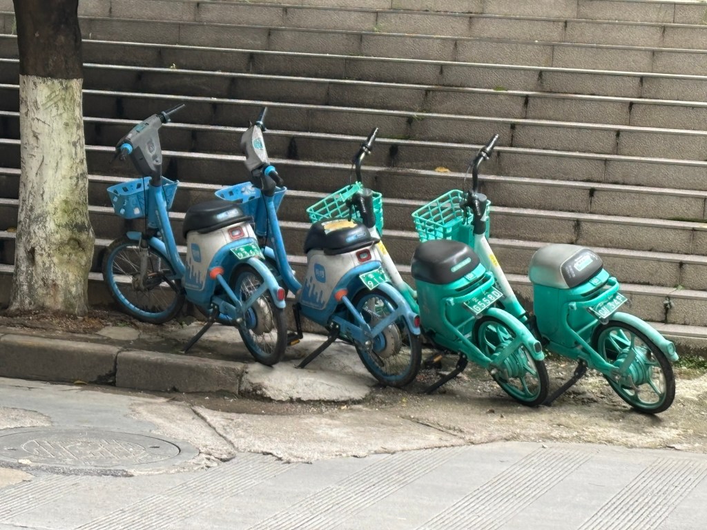 Vier kompakte E-Scooter mit Pedalen stehen nebeneinander am Straßenrand vor einer breiten Steintreppe. Sie sind türkis und hellblau lackiert, haben kleine Vorderräder, breite Sitzbänke, hinten ein Kennzeichen und vorne Einkaufskörbe am Lenker. Alle stehen auf dem Seitenständer, der linke direkt neben einem Baum.