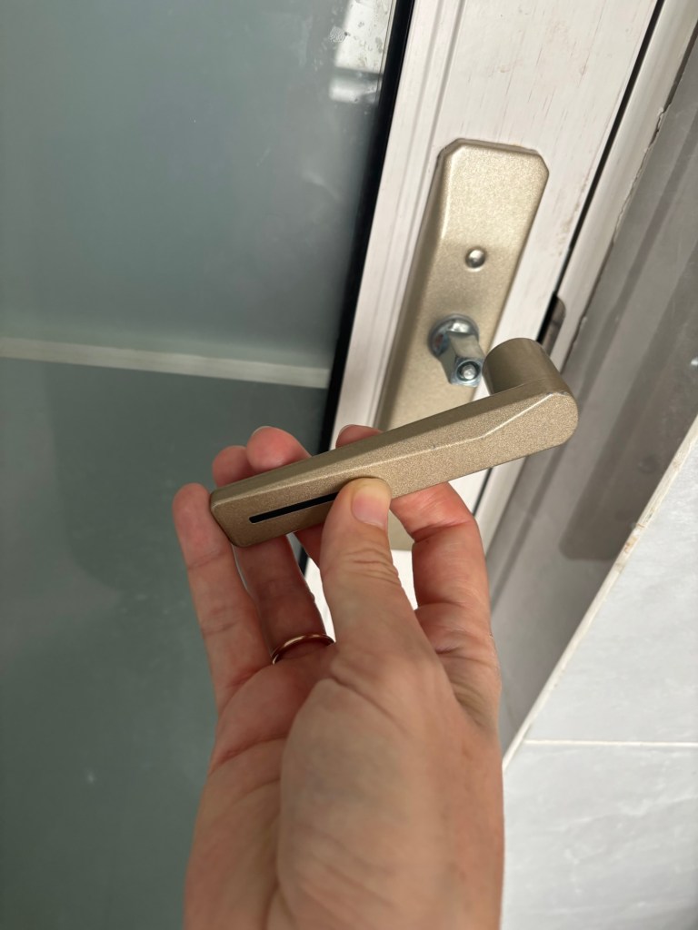 Die Tür mit der fehlenden Klinke im Hintergrund. Davor eine Hand mit der passenden Klinke. Es liegt nahe, dass als nächstes die Klinke auf die Schraube gesteckt wird.