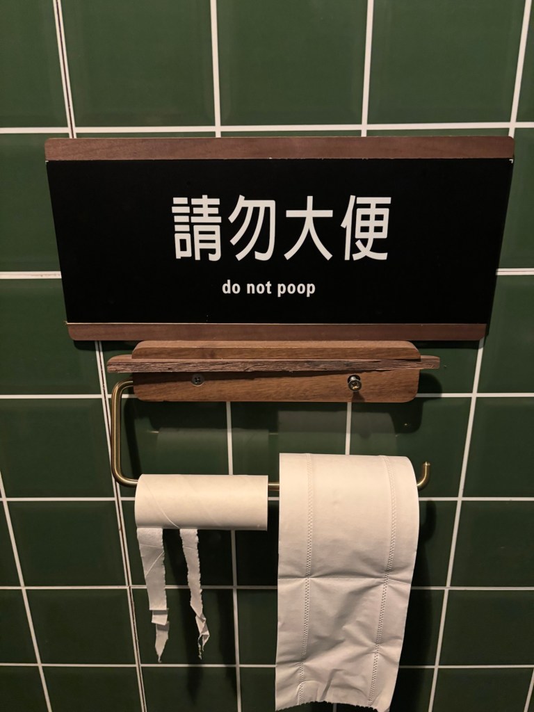 An einer dunkelgrün gefliesten Wand hängt ein Klorollenhalter mit Klopapier. Darüber ein Schild: „請勿大便“ „do not poop“