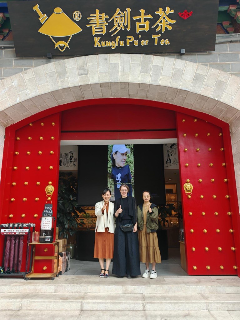 Drei Personen stehen vor einem Geschäft mit dem Schriftzug „Kungfu Pu’er Tea“ über einem großen roten Eingangstor mit goldenen Knöpfen. Sie lächeln und zeigen den Daumen nach oben. Links neben dem Eingang stehen rote Regenschirme in einem Ständer.