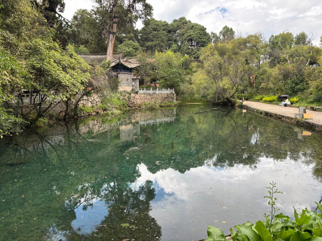 Ein Teich, in dessen ruhigem Wasser sich die Bäume und der Himmel spiegeln, ganz hinten unauffällig in braun gehalten ein Gebäude zwischen Bäumen