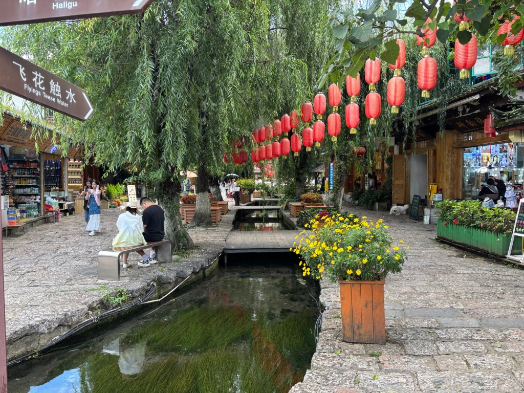 Kopfsteinpflaster, ein Kanal, kleine Brücken über den Kanal, rote Lampions sind zwischen Häusern und einer Trauerweide gespannt