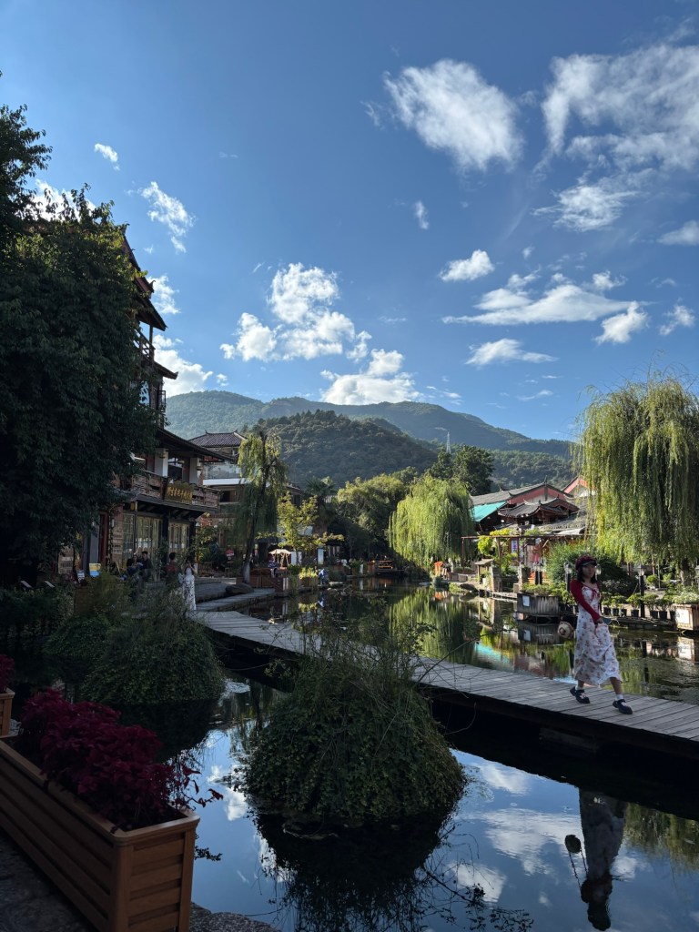 Eine größere Wasserfläche, gerahmt von Häusern, Bäumen, im Hintergrund Berge. Der Himmel ist blau mit nur einzelnen Wolken