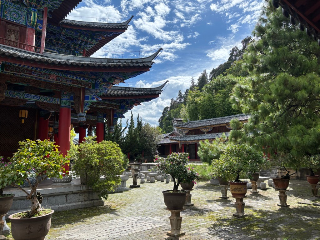 Bäume in Töpfen, links ein Tempel in traditioneller chinesischer Bauart mit roten Säulen, Bemalung in blauer Farbgebung und geschwungenen Dächern, in Hintergrund steigt ein Berg an