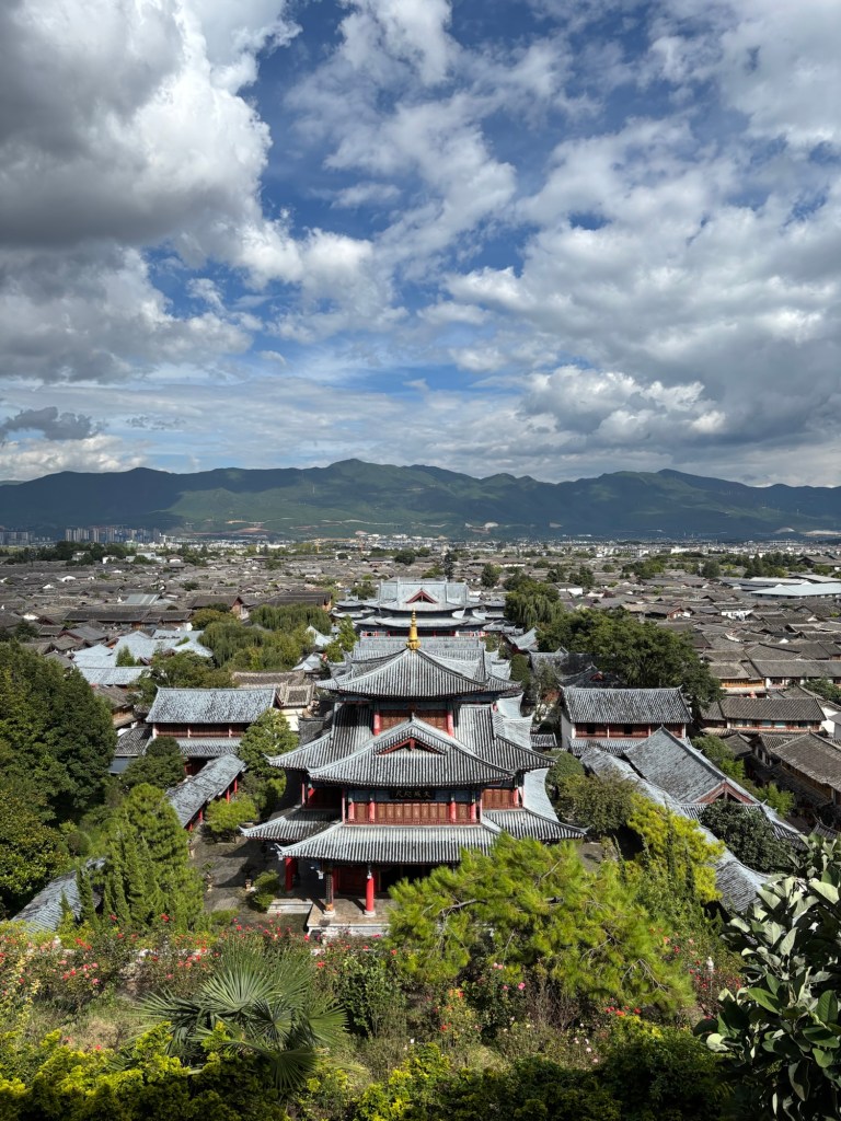 Blick von oben auf die Altstadt von Lijiang, die Häuser der Mu Familie sind in einer geraden Achse angeordnet und an den geschwungenen Dächern erkennbar. Im Hintergrund Berge und ein locker bewölkter Himmel.