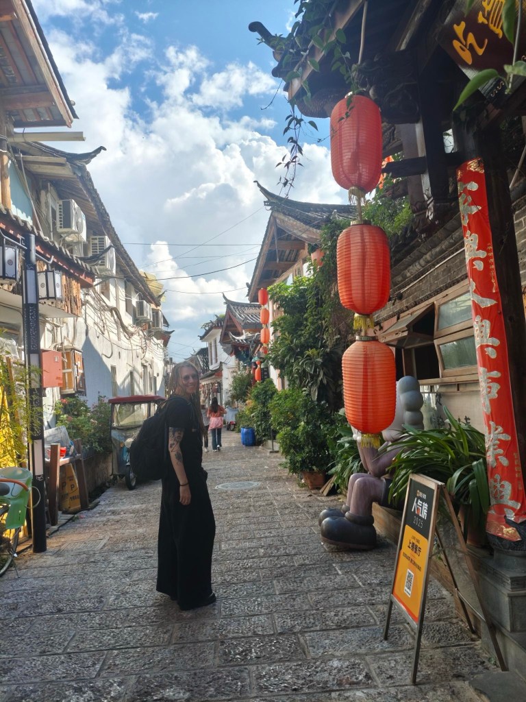 In einer von Lijiangs Gassen mit Kopfsteinpflaster schaue ich über meine Schulter in die Kamera und lache. Um mich Häuser mit viel grünen Pflanzen und roten Lampions