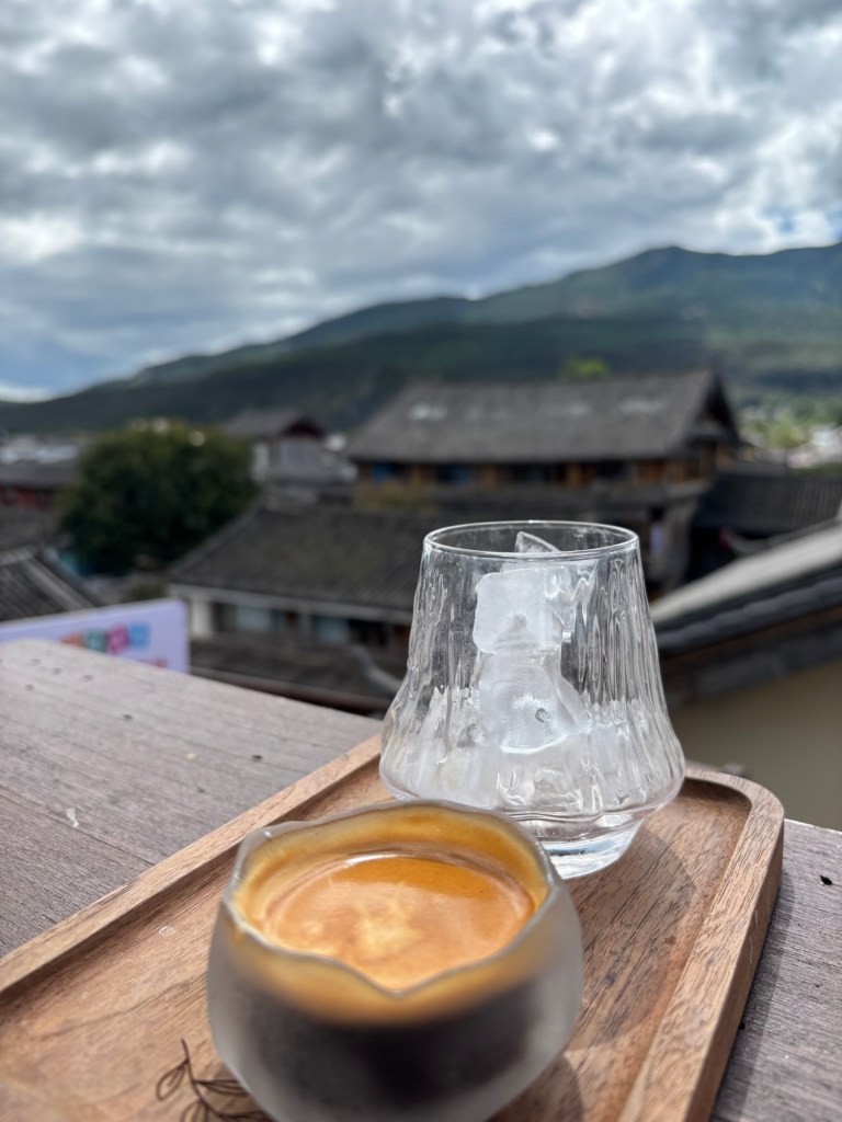 Auf einem Holztisch steht ein kleines Holztablett mit einem Espresso in einem wie eine Lotosblume geformten Glas und ein Glas mit Eiswürfeln. Im Hintergrund sind unscharf Dächer und Bergpanorama zu sehen