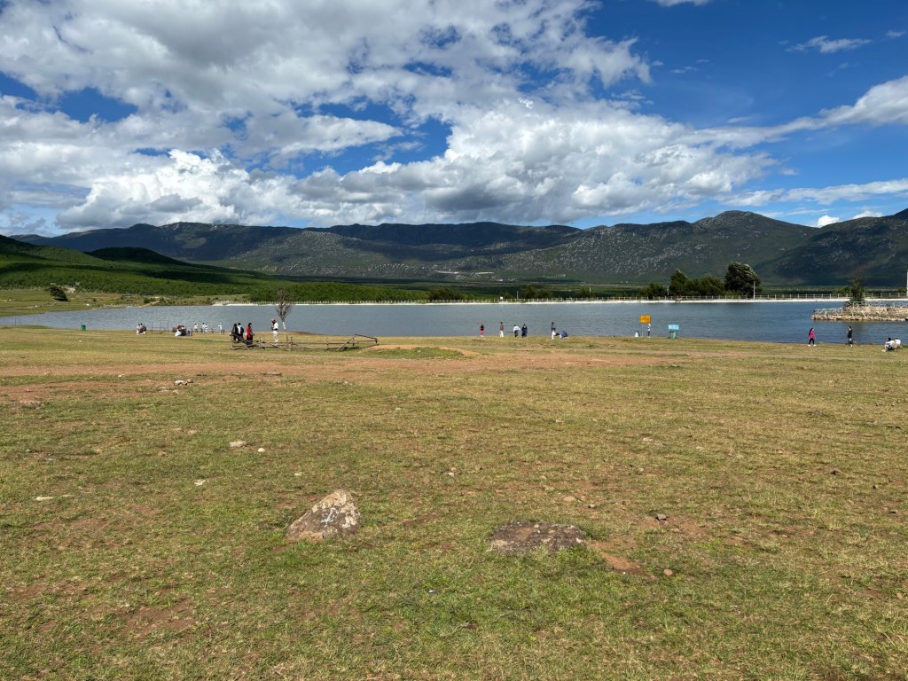 Berge, ein See, blauer Himmel mit wenigen weißen Wolken, Menschen stehen in kleinen Grüppchen zusammen