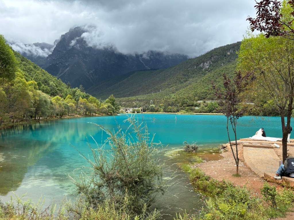 Leuchtend hellblaues Wasser, darum Berge und sehr tief hängende weiße Wolken