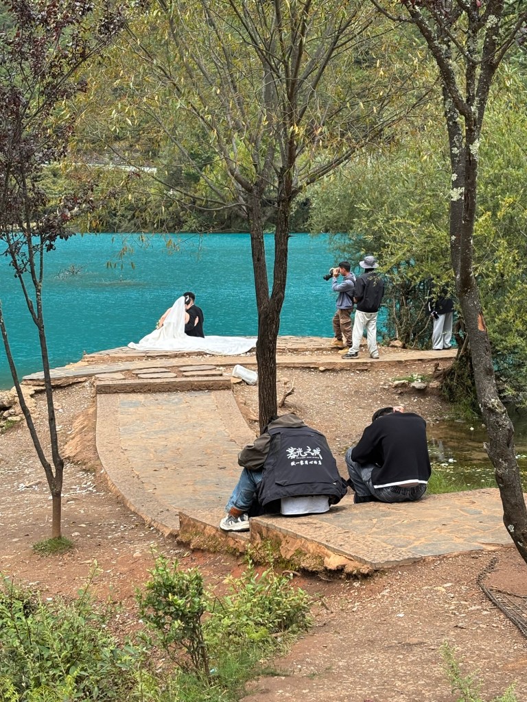 Auf einem Steg sitzt ein Hochzeitspaar, Gesicht zum See, das weiße Hochzeitskleid raumgreifend ausgebreitet. Die Braut blickt über die Schulter des neben ihr sitzenden Bräutigams in die Kamera