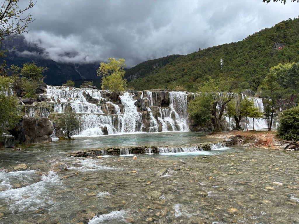 Ein breiter Wasserfall über mehrere Stufen, dahinter Berge und tiefe Wolken