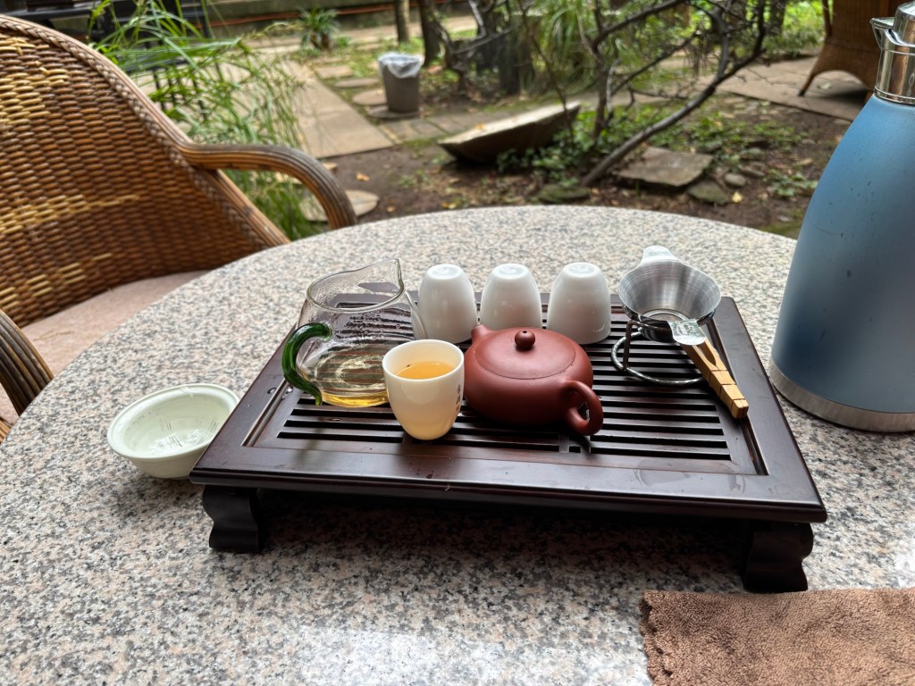Ein Marmortisch mit einem Holzbrett und Gitter, das das Ablaufen von Wasser erlaubt. Darauf eine tönerne braune kleine Teekanne, ein Gongfu Glas mit hellem Tee darin und eine kleine weiße Tasse, ebenfalls mit dem hellen Tee gefüllt. Im Hintergrund Pflanzen und Bäume