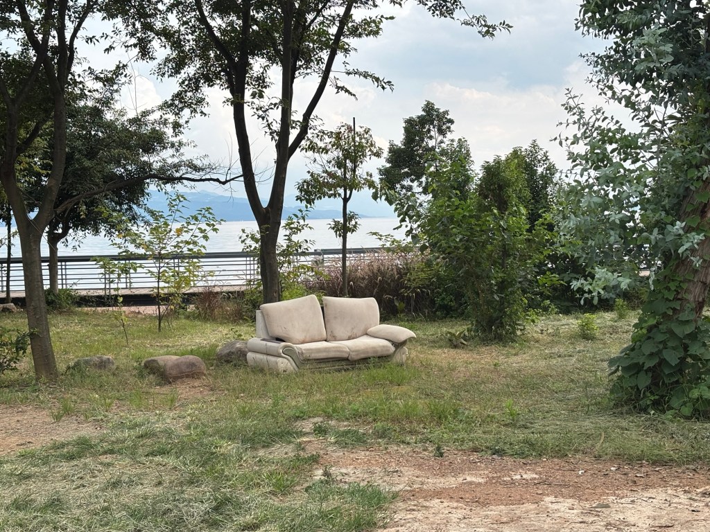 Eine wohl ehemals weiße, jetzt eher graue Zweisitzercouch steht unter freiem Himmel auf der Wiese