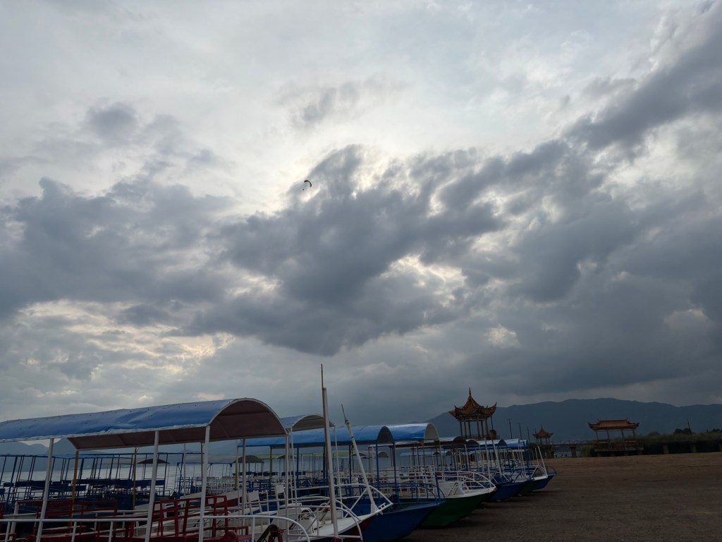 Am Strand liegen hochgezogene Boote, es ist kein Mensch zu sehen, am Himmel einige graue Wolken, im Hintergrund ein kleines Gebäude mit chinesisch geschwungenem Dach