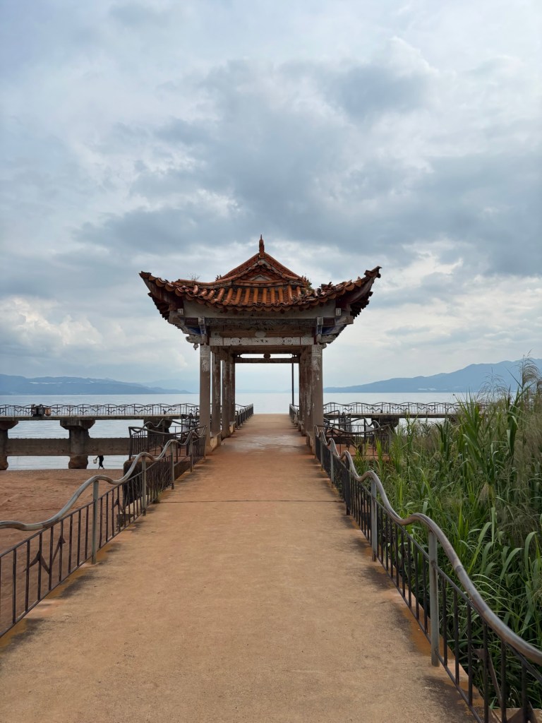 Ein etwas heruntergekommener Weg führt auf eine kleine Pagode zu, dahinter erstreckt sich der Fuxian See