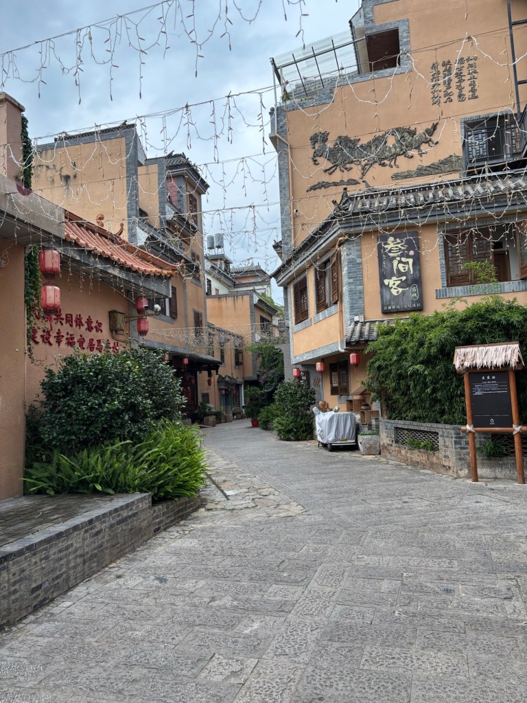 Eine verlassene kleine Gasse mit maximal dreigeschossigen kleinen Häusern in braun-beiger Farbgebung. An der rechten Hauswand sind chinesische Zeichen angebracht