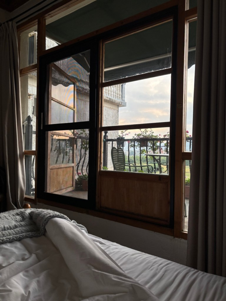 Blick vom Bett auf einen kleinen Balkon mit Blumen, dahinter der hellrosa eingefärbte Morgenhimmel