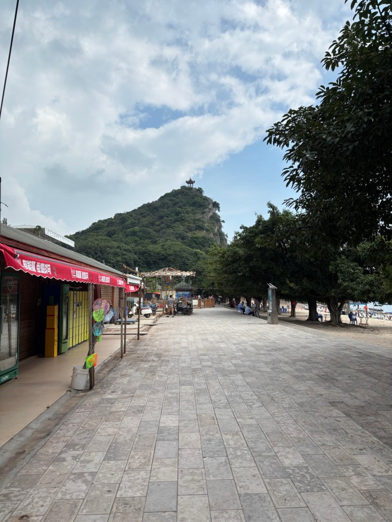 Eine Promenade mit Ladengeschäften läuft auf einen Berg zu, auf dessen Spitze eine Pagode zu sehen ist.