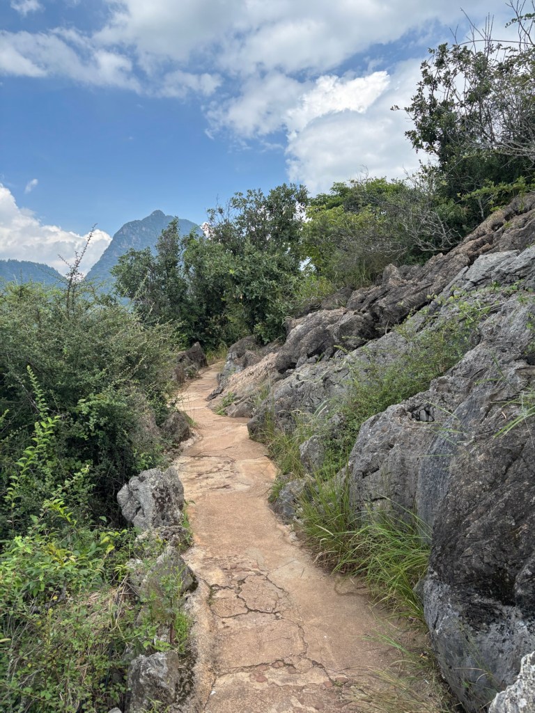 Ein schmaler gewundener brauner Weg, rechts begrenzt durch Felsen, links wird der abfallende Hang von Büschen gehalten