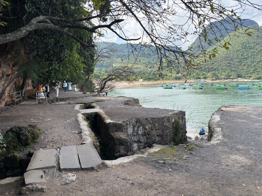 Gemauerte Promenade entlang des Sees mit einem Kiesweg entlang des Ufers. Bäume hängen über dem Weg. Im Hintergrund Boote vertäut auf dem See in Ufernähe.